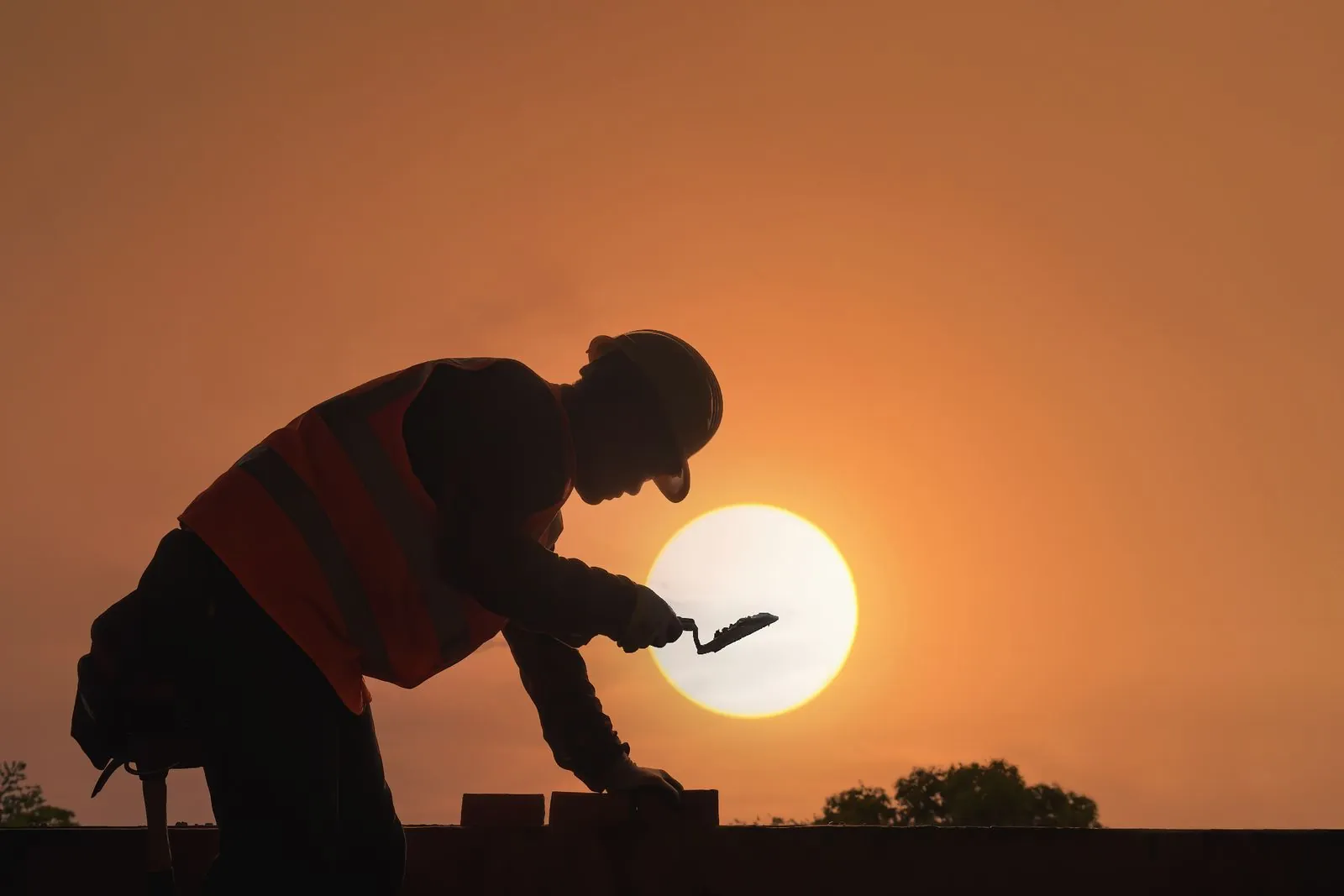 Silhouette of a bricklayer in a hard hat and hi-vis vest laying bricks as the sun sets behind him