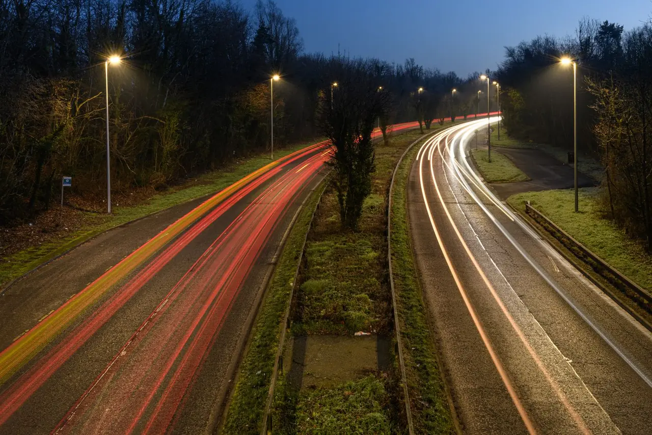 Evening traffic on a tree-lined dual carriageway at dusk, with headlights and tail lights streaking past under street lights