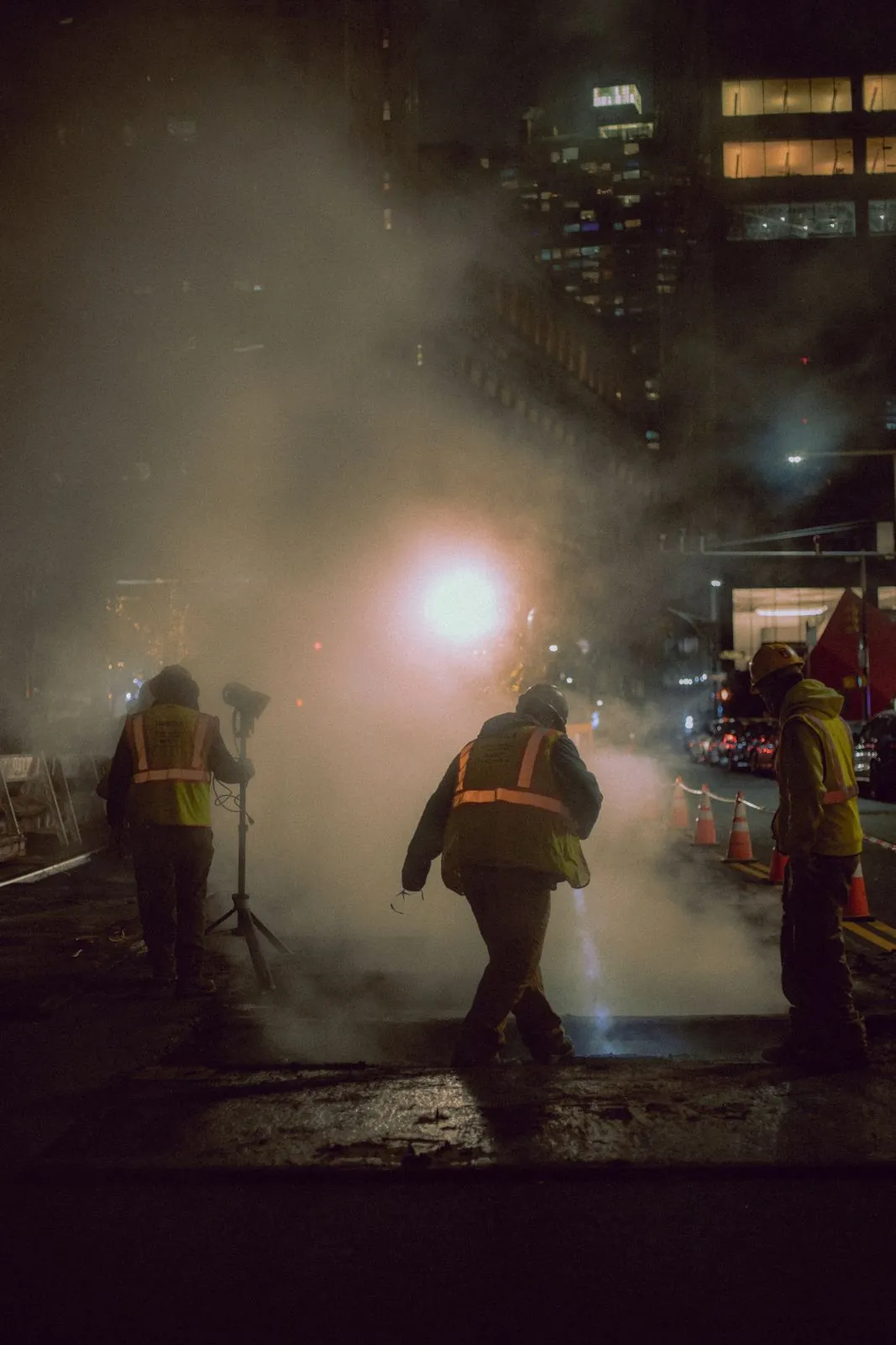 Road workers in hi-vis vests and hard hats working under a single work light at night, with traffic cones behind them